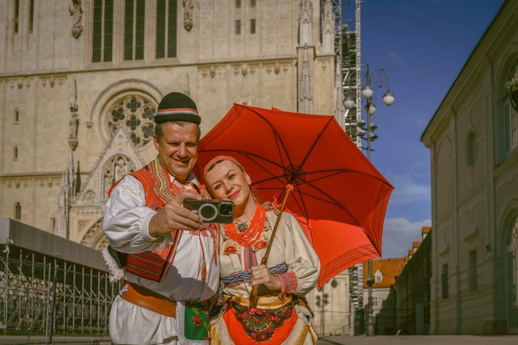 2 people dressed in folk attire holding red umbrella
